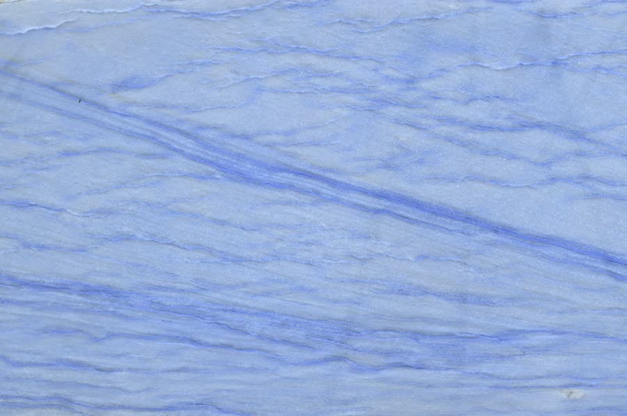 Close-up of a polished stone surface in soft cornflower blue and white, with fine horizontal layering and a pronounced deeper blue diagonal vein running from the upper left toward the right edge.