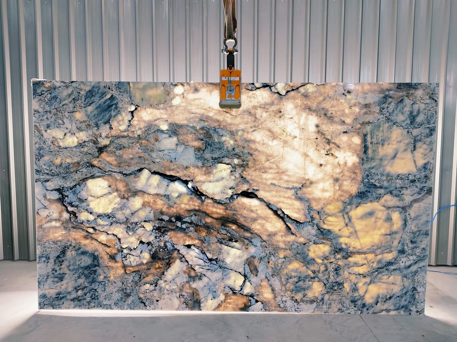 A large Blue Alpine slab lit from behind, hung from an overhead lifting clamp against a corrugated metal wall, with translucent amber and gold passages glowing through blue-grey and white mottled veining.