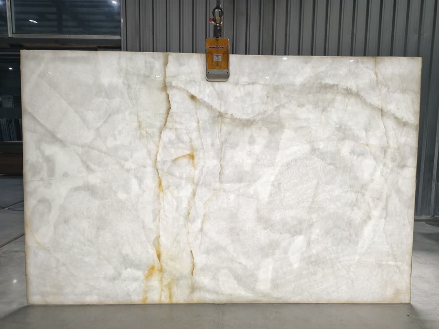 A full Crystallo Traslux slab held upright by a lifting clamp, near-white and translucent, with soft grey shading and thin rust-gold veins running down the left of centre.