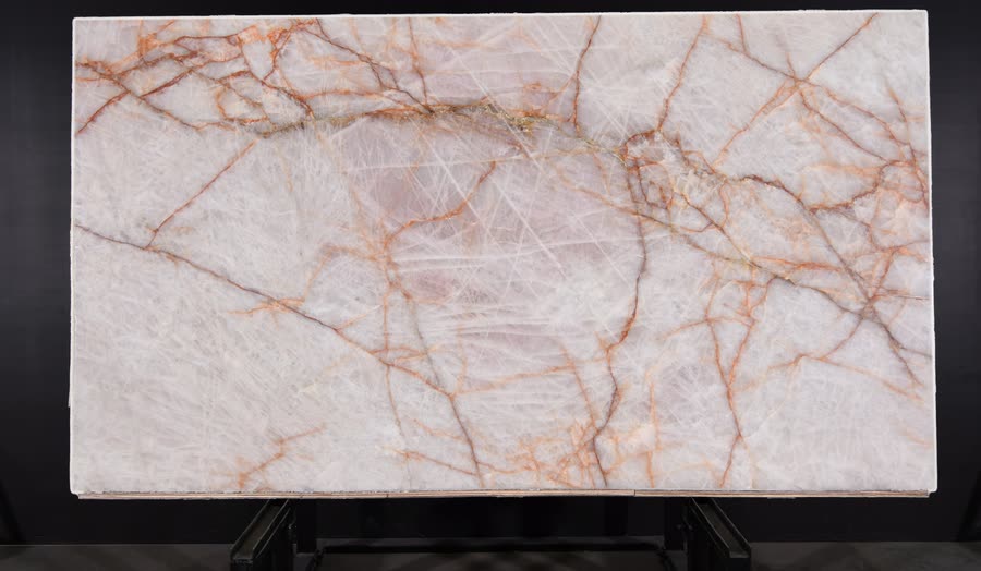 A full Rosso Crystallo slab on a dark metal easel against a black studio wall, translucent creamy white with a soft blush centre and a network of thin rust-orange veins.