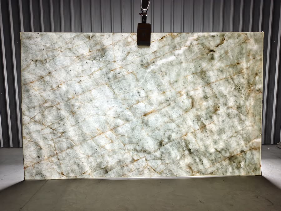 A large polished pale grey-green stone slab with white crystalline patches and rust-coloured seams, suspended upright by a lifting clamp against a corrugated metal wall on a concrete warehouse floor.