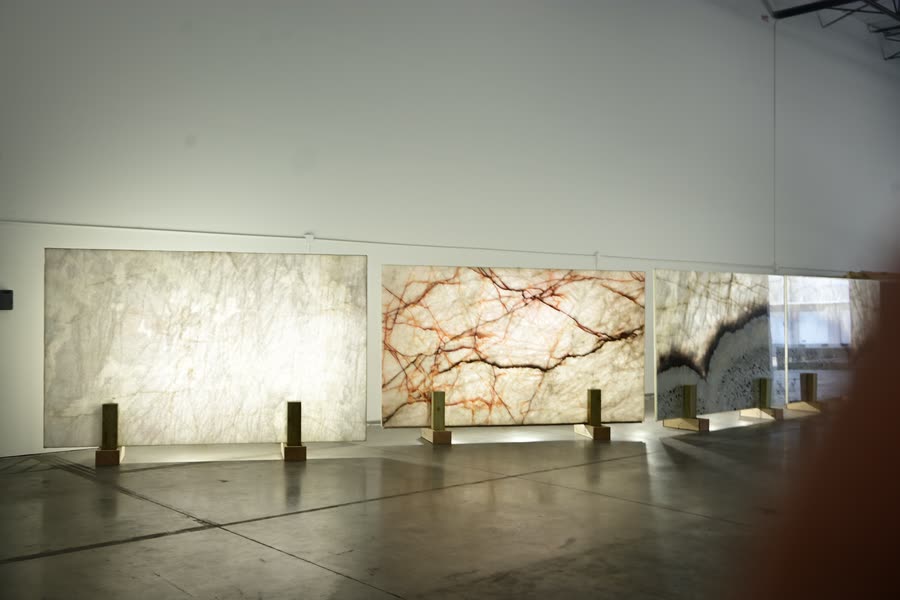 Three backlit stone panels on brass plinths against a white wall: the left panel glowing pale cream, the centre panel showing red and orange branching veins, the right panel banded in dark grey and white.