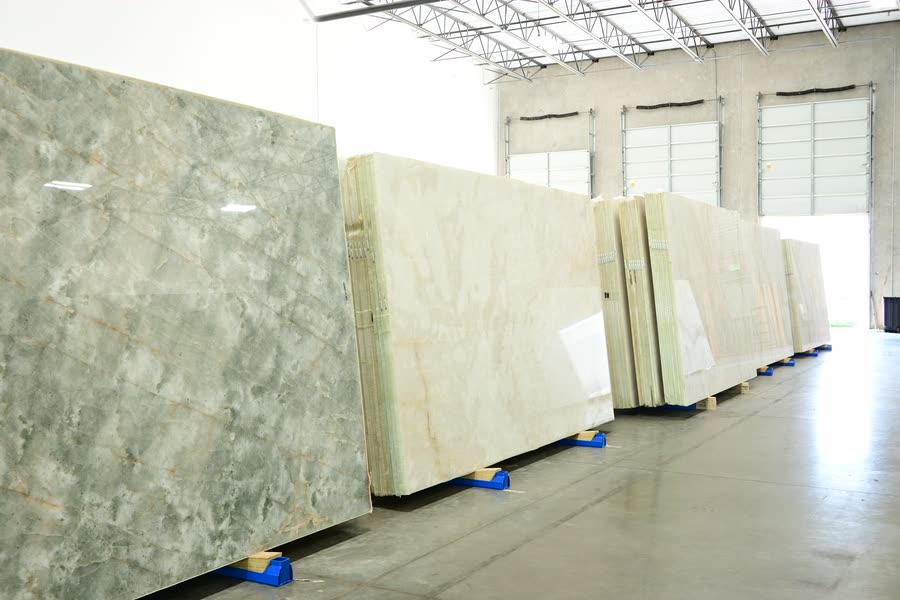 A line of stone slab bundles on blue plastic feet receding across a warehouse floor toward closed loading-bay doors, the nearest slab a large green-grey polished face catching the overhead lights.
