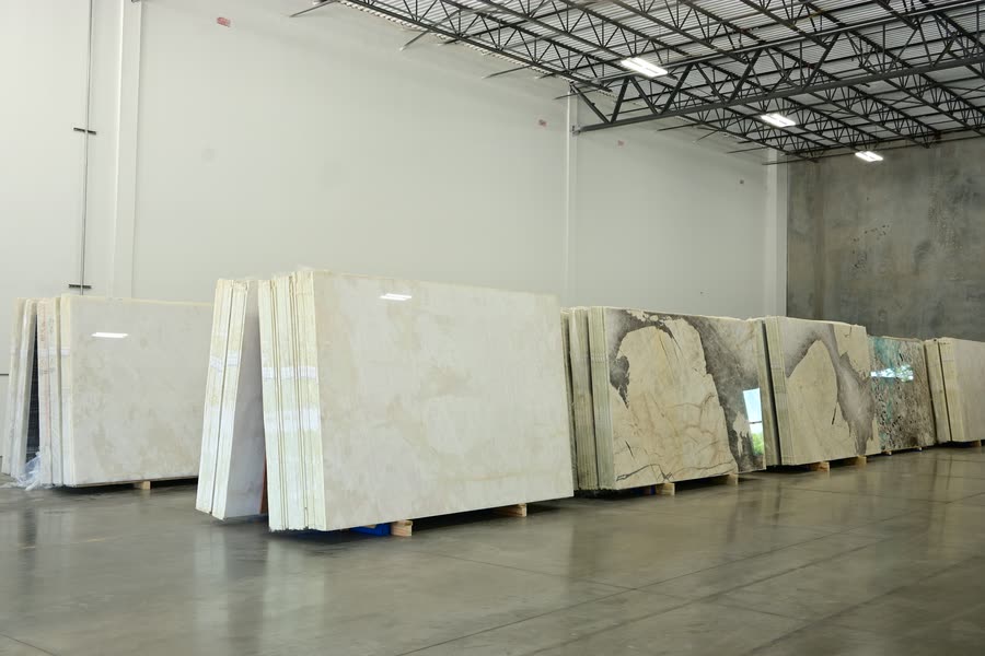 A row of stacked stone slab bundles resting on wooden pallets along the wall of a warehouse, receding to the right, with pale cream slabs in the foreground and darker veined and green-toned slabs further back on a polished concrete floor.