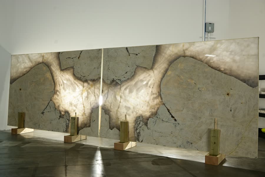A bookmatched pair of stone slabs standing side by side, their pale grey and cream veining mirrored across the central seam, resting on timber blocks on a dark polished floor.